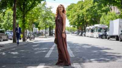 Chloé Lecareux wears a halter-neck dress in a rich brown satin fabric, featuring a flowing silhouette. The dress is complemented by a gold metallic collar detail. Hair is styled in loose waves. Accessories include a chunky bracelet with large, dark stones set in gold frames. A textured sequined brown handbag is carried, outside Stephane Rolland, during Paris Fashion Week - Haute Couture Fall/Winter 2025/2026, on July 08, 2025 in Paris, France